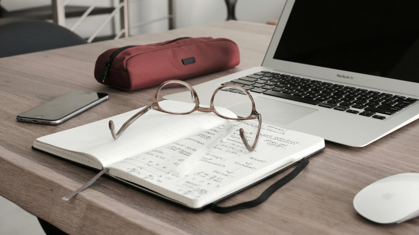 Laptop, notebook, glasses, and phone on a wooden desk.